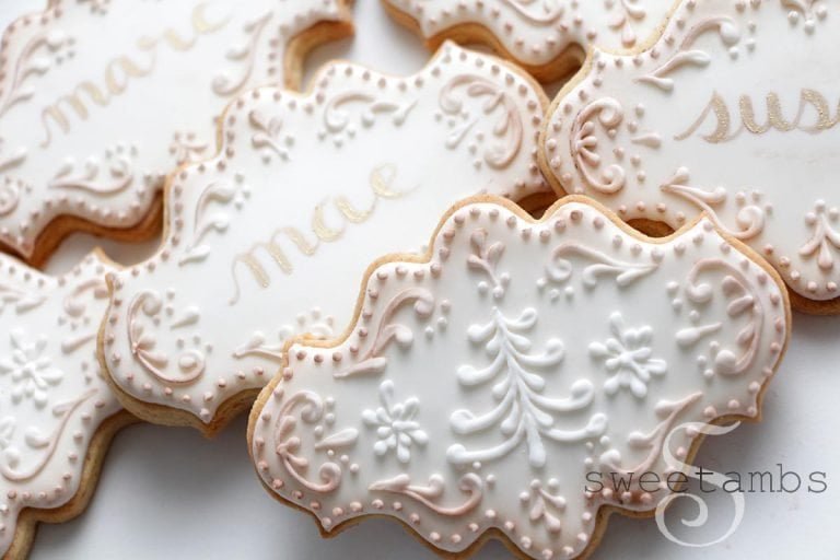 A pile of white sugar cookies with intricate patterns and the name mae and susan written on them in gold icing